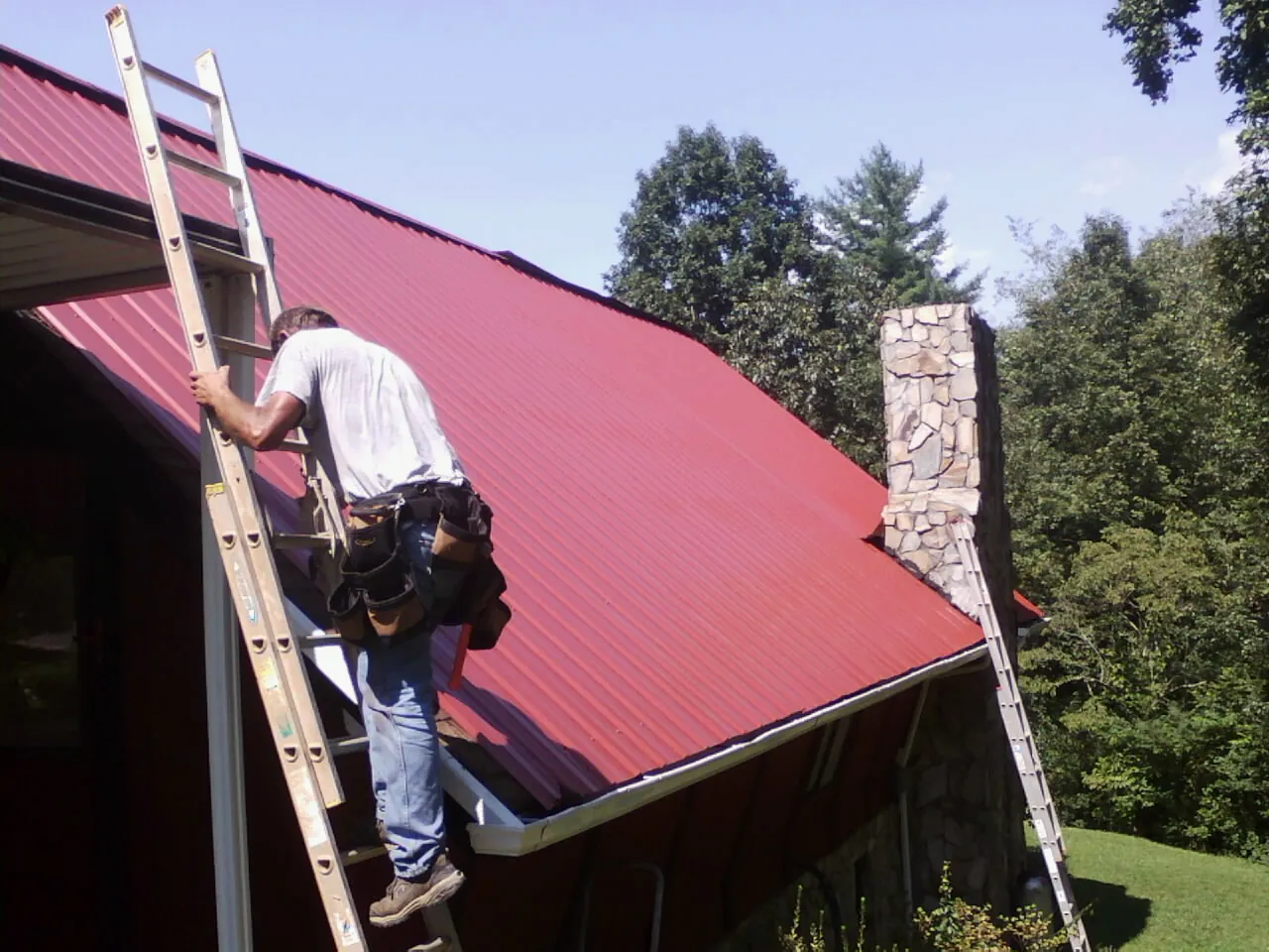 Roofer on a ladder beside a red metal roof and stone chimney — Soffit & Fascia Repair by Roofing Brothers in Moxahala